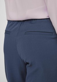 Gros plan d’une personne portant un pantalon bleu marine sur mesure avec des poches passepoilées à l’arrière et un chemisier rose clair rentré.