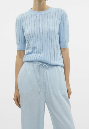 Persoon die een lichtblauwe gebreide top met korte mouw en bijpassende losse broek met trekkoord in de taille draagt, staand tegen een witte achtergrond.