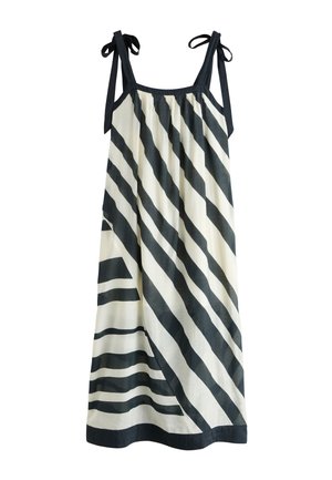 Joven mujer está de pie en un acantilado rocoso junto al mar, con un vestido largo a rayas blancas y negras, collar de conchas y sandalias negras.
