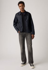Denim jacket in dark blue with light stitching, two front pockets, paired with gray jeans and black sneakers with white soles.