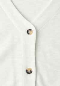Cardigan en maille blanche avec deux boutons ronds brillants en écaille fixés près de l'encolure en V.