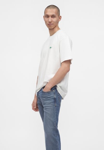 Homme aux cheveux courts portant un t-shirt blanc avec un petit logo et un jean bleu, debout avec une main dans la poche sur un fond uni.