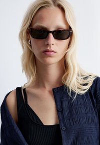 Schildpatt-Sonnenbrille mit dunklen Gläsern, getragen mit einem strukturierten, marineblauen Hemd, das über einem schwarzen Tanktop liegt.