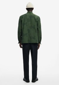 Green camouflage jacket made of fabric with a smooth texture, featuring a relaxed fit and long sleeves, paired with black trousers and shoes.