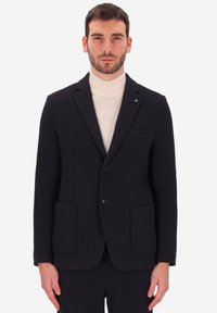 Blazer blu navy con texture, revers a punta, due tasche frontali e un dettaglio con taschino sul petto. Indossato sopra un dolcevita crema.