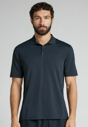 Man met krullend haar en baard, gekleed in een zwart poloshirt met korte mouwen, kijkt recht vooruit tegen een effen lichtgrijze achtergrond.