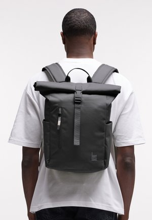 Mand iført hvid t-shirt og sorte bukser med en sort rulle-top rygsæk med frontlynlås og sidelommer.