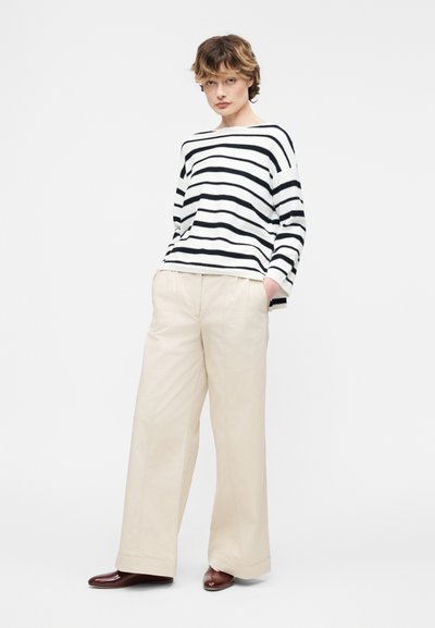 Άτομο που φοράει μακρυμάνικη μπλούζα με λευκές και μαύρες ρίγες, μπεζ παντελόνι φαρδύ στο πόδι και καφέ παπούτσια, στέκεται με τα χέρια στις τσέπες μπροστά σε απλή φόντο.