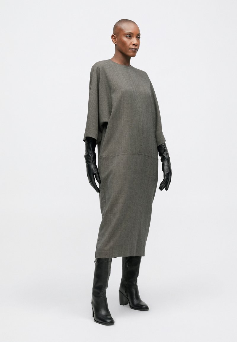 Robe en tricot longue et grise avec un col haut et des manches chauve-souris. Associée à des gants en cuir noirs ajustés et des bottes noires à talons montantes jusqu'aux genoux.