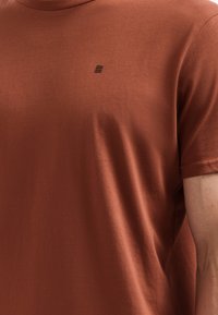Roestoranje T-shirt van zachte stof, met een ronde halslijn en een klein donker logo-detail op de borst. Korte mouwen.