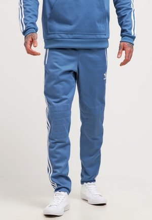 Man wearing blue Adidas track pants with white stripes and white sneakers, standing against a plain background.