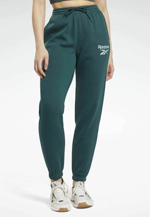 Personne portant un pantalon de jogging Reebok vert foncé et des baskets blanches, debout devant un fond clair uni.