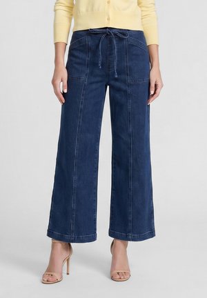 Pantaloni in denim blu scuro a gamba larga con coulisse frontale, indossati con sandali beige con tacco alto e punta aperta e un cardigan giallo chiaro.