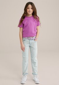 Lila kortärmad t-shirt med ett solemblem, kombinerad med ljusblå slitna jeans och ett mönstrat rosa bälte. Vita sneakers fulländar looken.