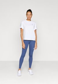 Biele športové tričko s krátkym rukávom v kombinácii s modrými legínami. Tričko má logo; legíny majú logo a hladkú textúru. Biele tenisky.