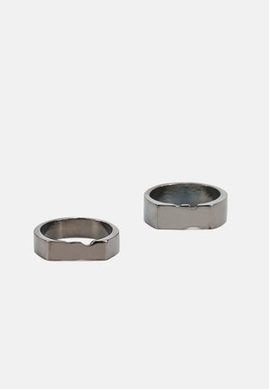 Dois anéis metálicos com acabamento escovado. Ambos têm um design robusto e uma forma quadrada, apresentando um entalhe de um lado para detalhe.