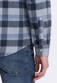Camisa de franela a cuadros azules que presenta tonos variados de azul con acentos blancos. Con puños de botón y un dobladillo curvado. Combinada con jeans de denim oscuro.
