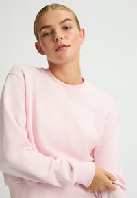 Felpa rosa con scollo rotondo, maniche lunghe, polsini e orlo a coste, realizzata in tessuto morbido, con un piccolo logo sul petto.