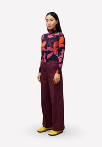 Langärmliges Turtleneck-Oberteil mit buntem Blumenmuster; weite bordeauxrote Cordhosen; gelbe Schuhe mit schwarzen Sohlen.