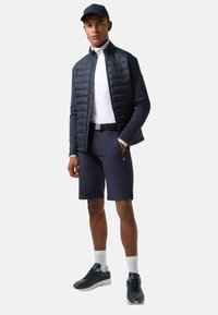 Marineblå pufferjakke over en hvid polo, parret med marineblå shorts og mørke sneakers. Hvide sokker og en matchende kasket fuldender outfittet.