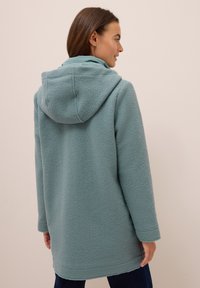 Vaaleansininen fleece-takki, jossa on rento istuvuus, pitkät hihat ja helma, joka yltää juuri reiden yläpuolelle. Pehmeää, teksturoitua kangasta.