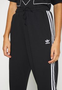 Svarta träningsbyxor med vita sidosträck, elastisk midja, dragsko, två sidofickor och Adidas-logotyp på vänster lår. Slät tygtextur.