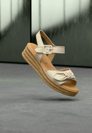 Beige strappy sandal with a knotted detail, adjustable ankle buckle, and a stacked wedge sole featuring a textured rubber base.