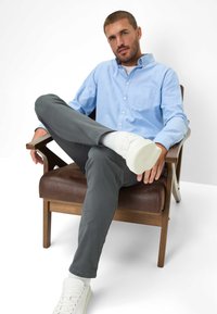 Chemise boutonnée bleu clair, pantalon gris, baskets blanches. Le modèle est assis sur une chaise en bois, les jambes croisées, avec un détail de poche sur la chemise.
