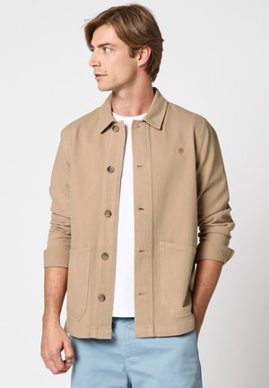 Homme portant une veste beige boutonnée avec poches par-dessus une chemise blanche et un pantalon bleu clair, regardant vers la gauche.