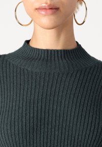 Maglione a coste di un verde scuro, con collo alto. Texture liscia e design aderente, abbinato a grandi orecchini a cerchio dorati.