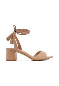 Sandalias beige de tacón abierto con tacón grueso, diseño con lazo en el tobillo y tiras planas. Textura de cuero suave con detalles de borlas.