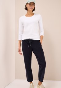 Mujer con una blusa blanca de manga larga, pantalones negros con cordón, zapatillas beige y una gorra verde claro, de pie contra una pared neutra.