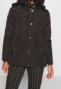 Svart vadderad jacka med huva, utrustad med guldfärgad maskinvara, tryckknappar och två framfickor. Strukturerat tyg med diamantmönster.