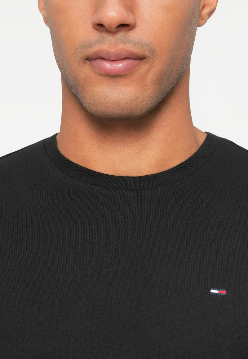 Black crew-neck t-shirt made of smooth fabric, featuring a small embroidered logo with red, white, and blue colors on the left chest.