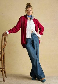 Red plaid shirt layered over a white blouse with lace detailing, paired with flared blue denim jeans and teal high-heeled shoes. Wood chair background.