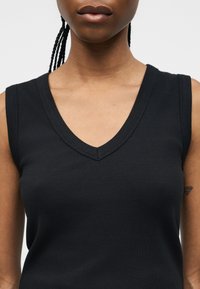 Γυναίκα που φοράει ένα μαύρο ριμπ πλεκτό αμάνικο τοπ με V-λαιμόκοψη, με πλεξούδες ορατές πίσω και ένα μικρό τατουάζ στον πάνω βραχίονα.