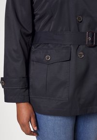 Chaqueta impermeable en color marino, con cintura ceñida, dos bolsillos frontales y detalles de botones. Textura suave con costuras decorativas.