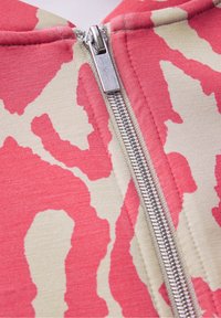 Dragkedja hoodie med en beige tyg och ett rosa abstrakt mönster. Metalldragkedjadetalj löper ner i mitten. Mjuk, texturerad material.