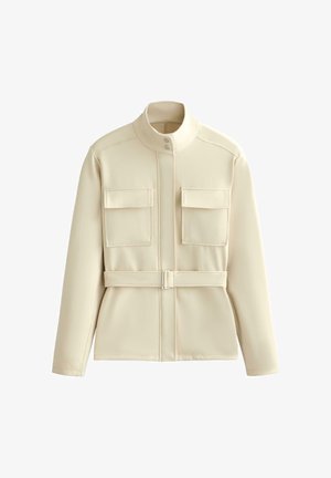 Bézs hosszú ujjú dzseki álló gallérral, két első zsebfedővel és hozzáillő övvel a deréknál, fehér háttéren bemutatva.