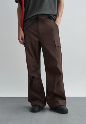 Calças cargo castanhas feitas de um tecido resistente, com um corte descontraído, vários bolsos e costuras discretas. Calçado preto visível.