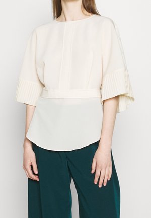 Kobieta w luźnej białej bluzce z szerokimi plisowanymi rękawami i ciemnozielonych szerokich spodniach, stojąca na prostym białym tle.