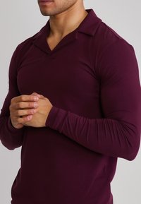 Bărbat purtând o cămașă strâmtă cu mâneci lungi, de culoare mov închis, cu guler deschis, cu mâinile încleștate la piept, pe un fundal simplu și deschis la culoare.