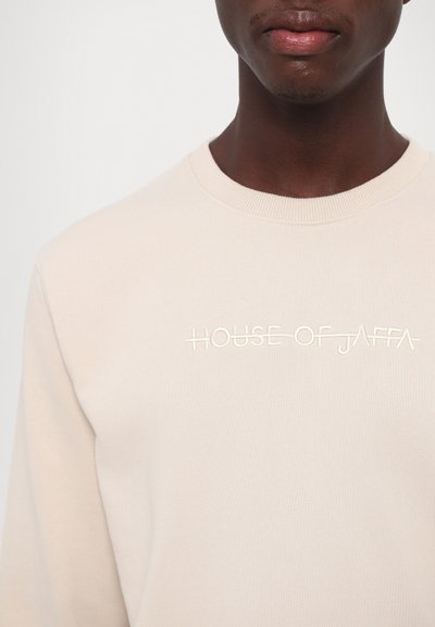 Beige genser med rund hals, med brodert tekst "HOUSE OF JAFFA" i kontrasterende hvitt, laget av mykt stoff.