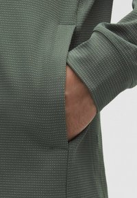 Zelená textúrovaná látka s malými bielymi bodkami; dlhé rukávy a bočná vrecko na úrovni pása. Ozdobené jemným švom.