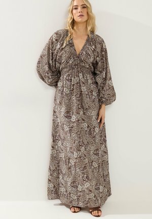 Mujer vestida con un vestido largo marrón con estampado de cachemira y mangas largas abullonadas, y sandalias, de pie contra una pared blanca.