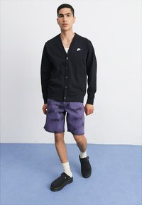Svart knäppbar cardigan, lila tie-dye shorts, vita ankelsockor och svarta träskor. Minimalistisk design med Nike-logotyp på cardiganen.