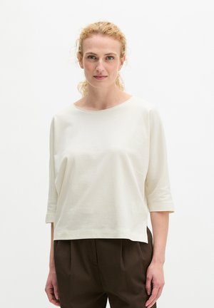 Femme aux cheveux blonds bouclés portant un haut blanc ample à manches trois-quarts et un pantalon marron foncé, debout devant un fond clair uni.