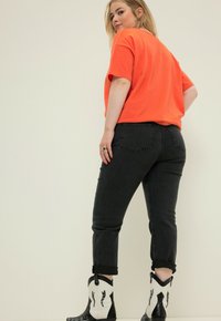 Jeans in denim neri con un design aderente e risvolti, abbinati a una t-shirt arancione oversize. Stivali da cowboy bianchi con dettagli neri.