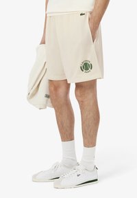 Shorts en maille beige avec le logo Lacoste Paris en vert et un graphique de tennis ; taille élastique ; poches latérales ; associés à des baskets blanches et des chaussettes.
