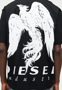 Μαύρο πουκάμισο με μεγάλο λευκό γραφικό φοίνικα και μερικό κείμενο "DIESEL industry" στην πλάτη, φορεμένο από άτομο με σκουρόχρωμο δέρμα.